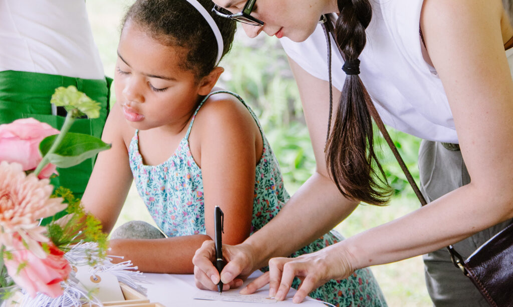 A young girl and a woman lean over a table as they write their wishes on small tags