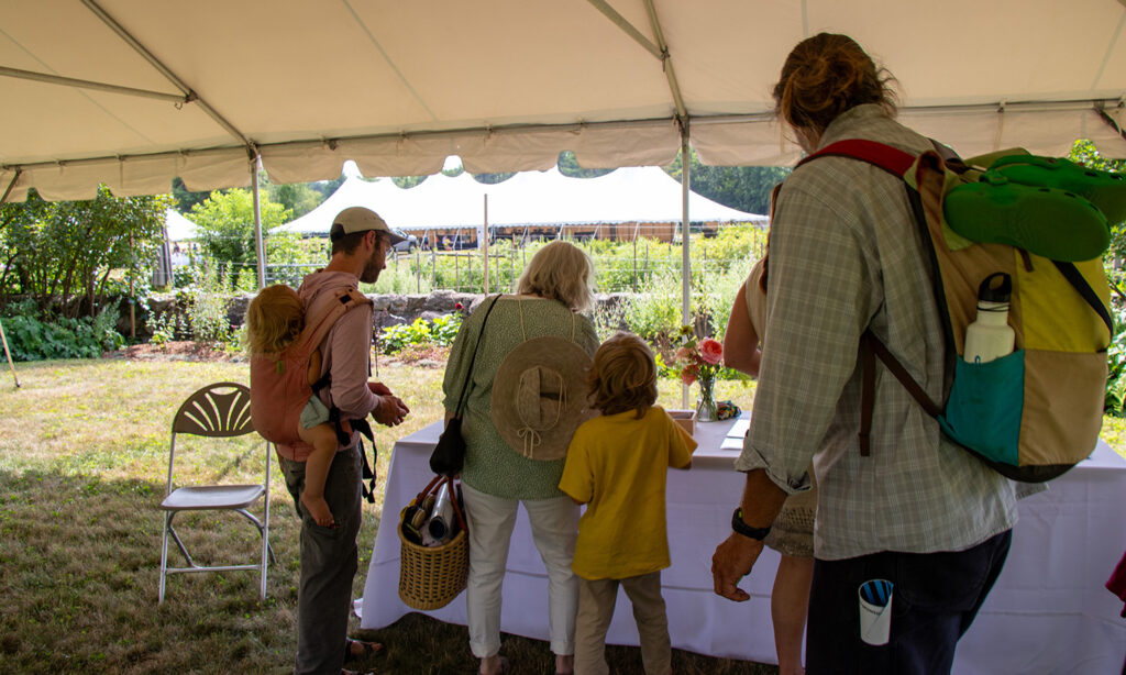 a small group gather under a white tent. They are gathering materials for an activity from a table