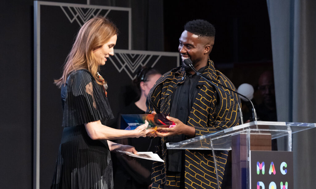 Two people stand, facing one another on a stage. One hands a trophy to the other.