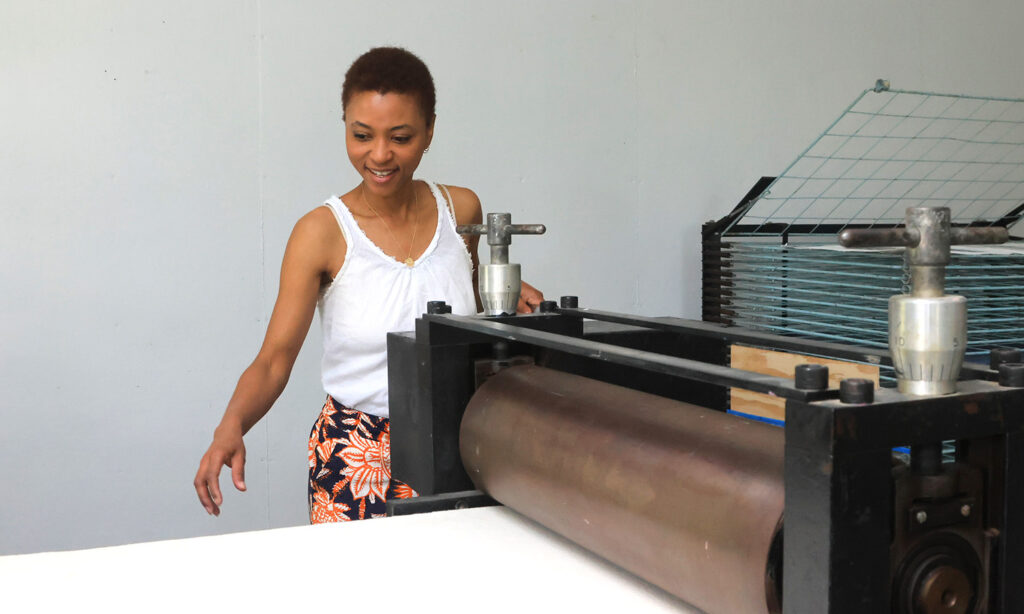 A woman, wearing a white tank top, floral printed pants, and sporting short hair, stands behind a printing press, gazing toward the press and operating the machine