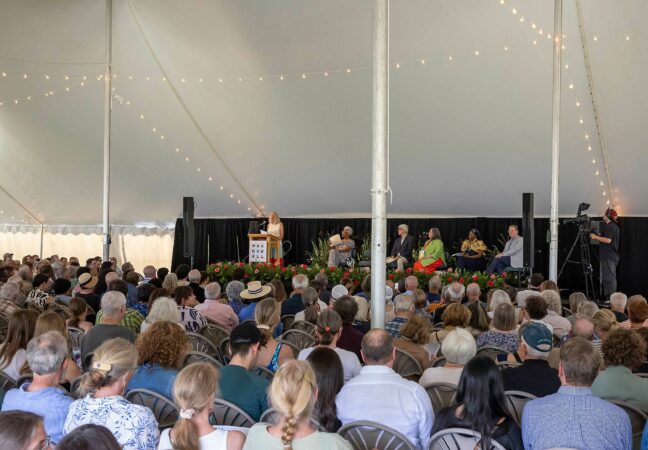 A large crowd of people sita under a tall tent listening to a speaker on stage in the background. The dais has five other people seated on it.