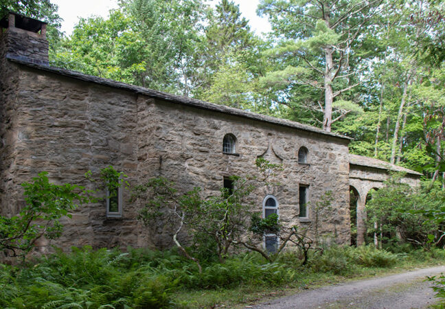 A granite stone building in a dark forest, next to a gravel road.