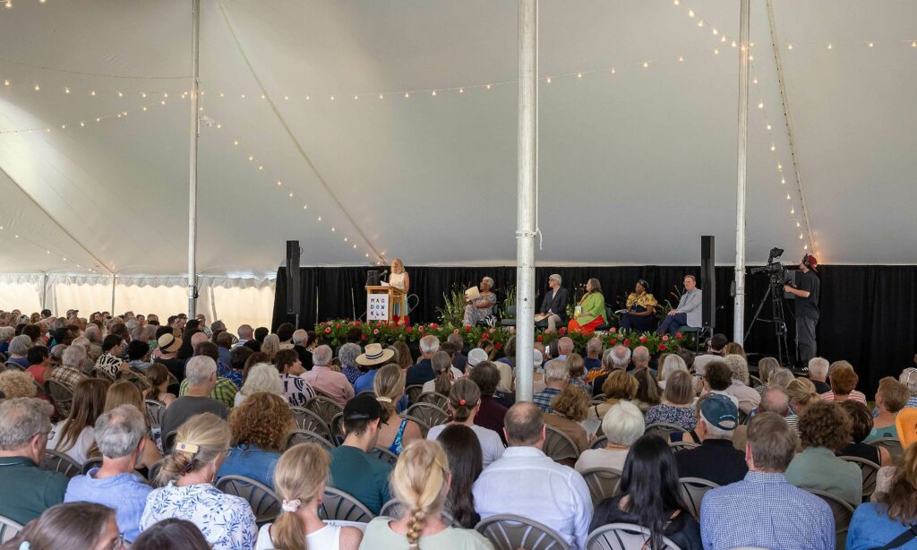 A large crowd of people sita under a tall tent listening to a speaker on stage in the background. The dais has five other people seated on it.