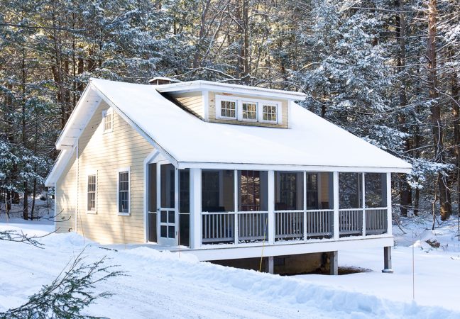 New Jersey Studio in January surrounded by snow. The yellow building is nestled against a dense forest that is dusted with snow. The sun, shining through the forest canopy, casts speckled light on the studio.