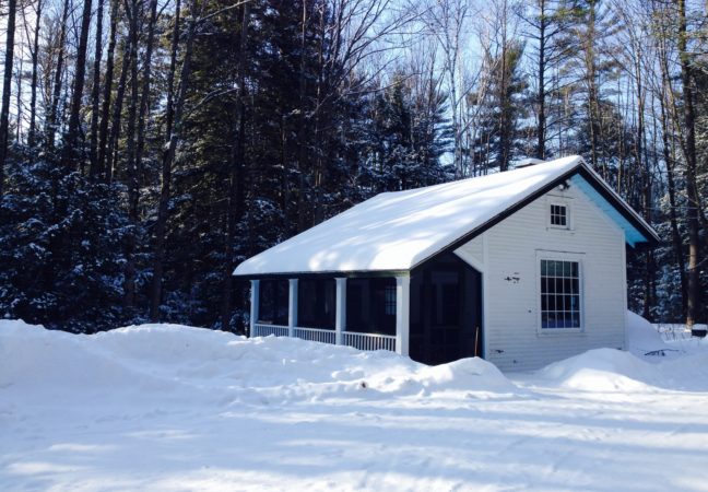 Monday Music Studio in winter surrounded by snow. The small white building, set in a clearing of a dense forest, is blanketed in white.