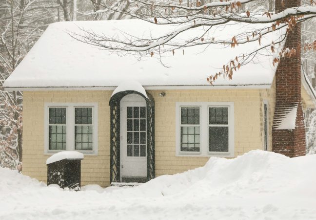 Mansfield Studio in winter surrounded by deep snow. The small yellow building, nestled into a dense forest, is blanketed in white.