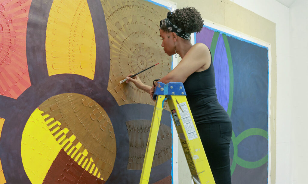 A woman stands on a ladder while painting on a massive canvas