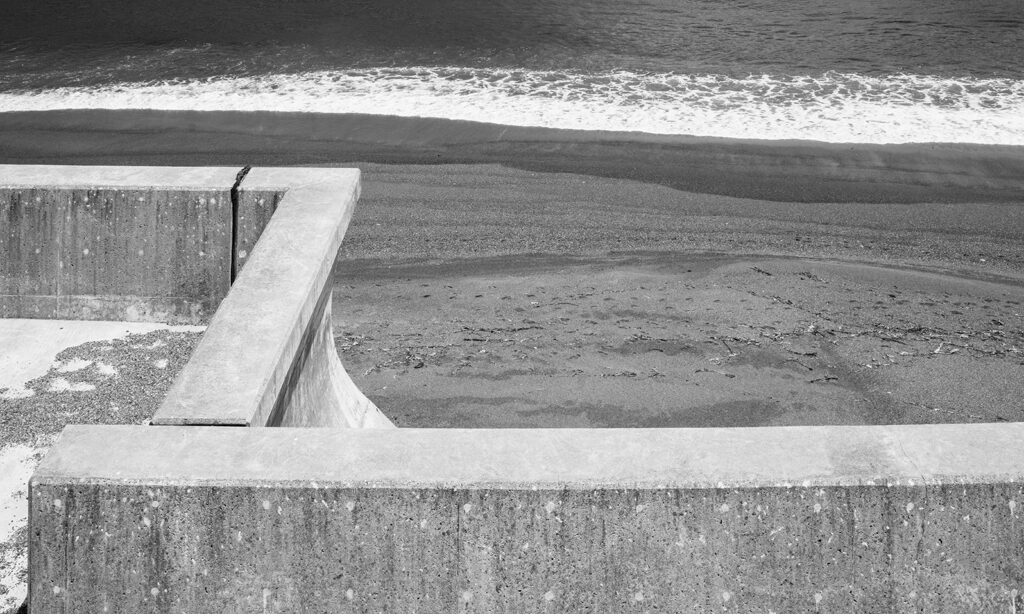 A black and white photo of concrete walls overlooking waves breaking on a beach 100 yards in the distance.